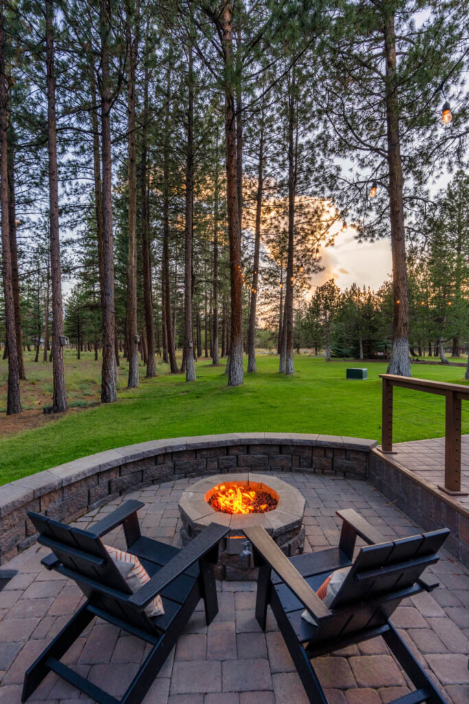 Backyard paver patio with fire pit and Adirondack chairs among tall pines at dusk