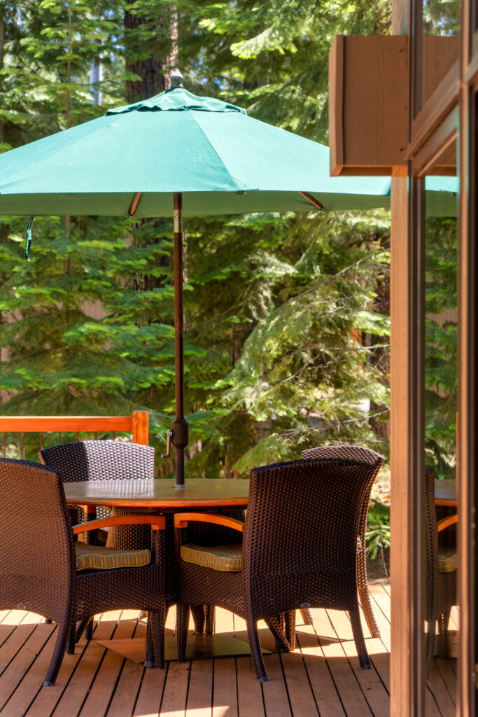 Shaded deck dining under a green umbrella surrounded by tall evergreens